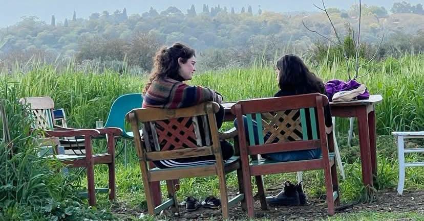 שתי נשים יושבות ומשוחחות על כיסאות עץ ליד שולחן בחיק הטבע
